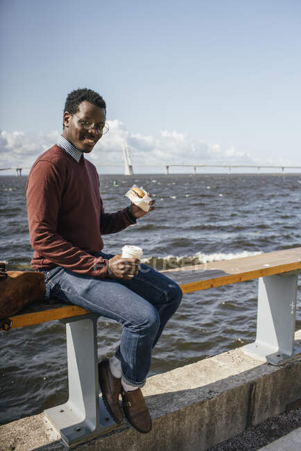 Молодой человек сидит на перилах у моря, ест гамбургер, пьет кофе — стоковое фото