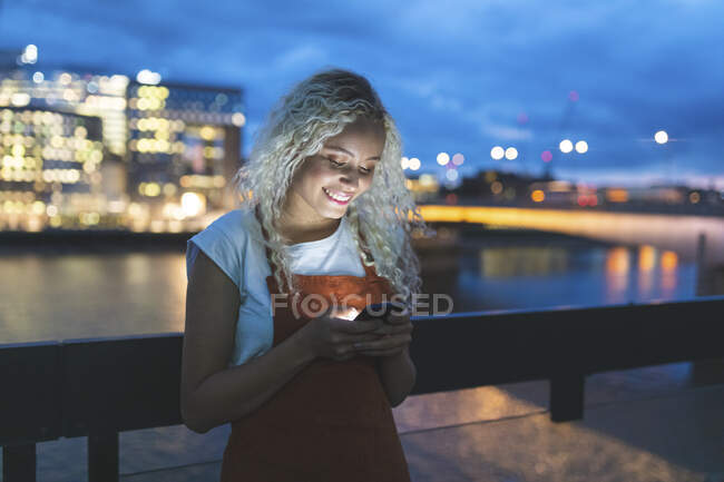 Молодая женщина в городе в сумерках смотрит на свой смартфон — стоковое фото