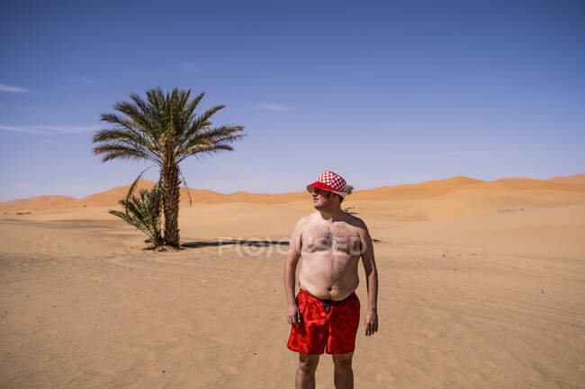 Homem de sobrepeso com calções de banho e chapéu em pé no deserto de Marrocos — Fotografia de Stock