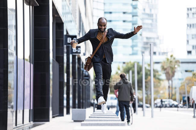 Jovem empresário com fones de ouvido e café para ir pulando de bollard para bollard — Fotografia de Stock