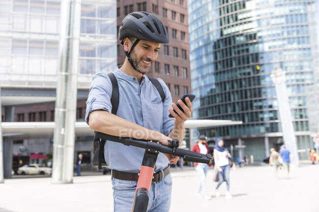 Усміхаючий бізнесмен дивиться на смартфон, який працює на електричному скутері в місті Берлін (Німеччина). — стокове фото