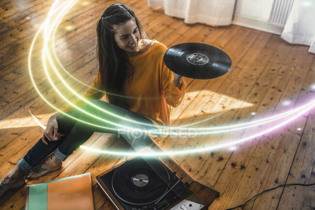 Young woman sitting on the floor at home with record and record player surrounded by a light trail — Stock Photo