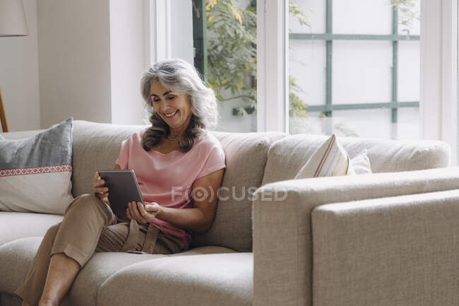 Посмішка дорослої жінки за допомогою таблетки сидить вдома на дивані. — стокове фото
