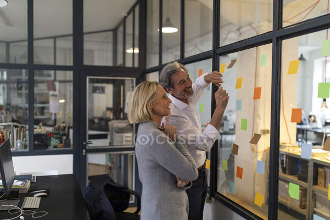 Glückliche Kollegen blicken im Büro auf klebrige Zettel an der Glasscheibe — Stockfoto