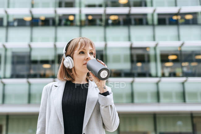 Жінка з навушниками, що п'ють з багаторазової пляшки в місті — стокове фото