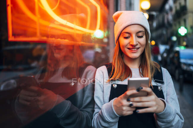 Lächelnde junge Frau, die nachts mit ihrem Smartphone neben Neonlicht steht — Stockfoto