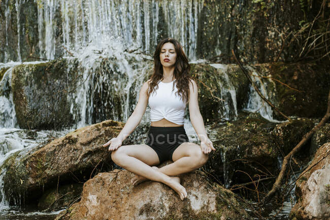 Junge Frau praktiziert Yoga auf einem Wasserfall, sitzend auf einem Felsen — Stockfoto