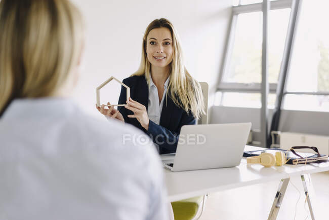 Smiling young businesswomen showing house model at desk in office — Stock Photo