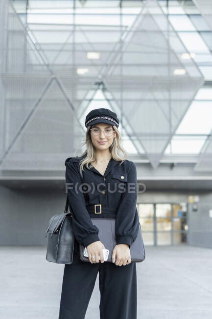 Giovane donna d'affari bionda con cappello da marinaio nero e borsa per laptop e smartphone, guardando la fotocamera — Foto stock
