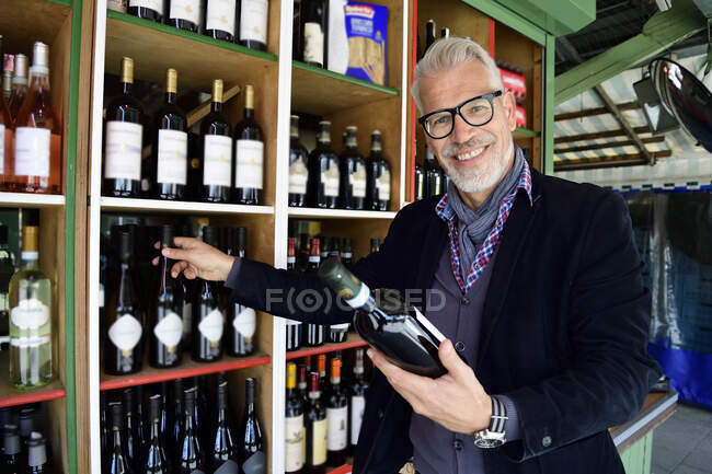 Retrato de hombre maduro sonriente eligiendo botella de vino en una tienda de vinos - foto de stock