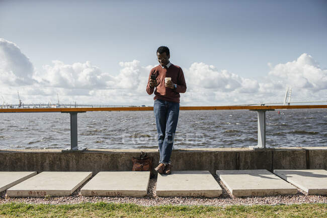 Giovane in piedi sul mare, tenendo il caffè, utilizzando lo smartphone — Foto stock