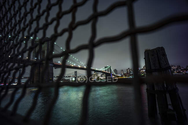 USA, New York, New York, Chainlink recinzione contro illuminato ponte di Brooklyn di notte — Foto stock