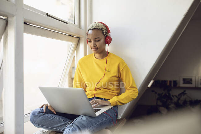 Jeune femme assise à la fenêtre à la maison avec ordinateur portable et écouteurs — Photo de stock