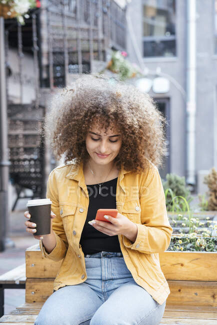 Портрет усміхненої дівчини-підлітка, яка сидить на лавці з кавою і дивиться на мобільний телефон. — стокове фото