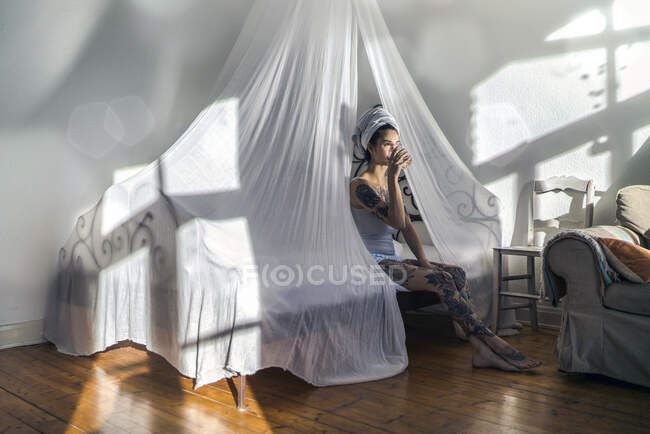Tatoué jeune femme boire un verre d'eau dans le lit à baldaquin — Photo de stock