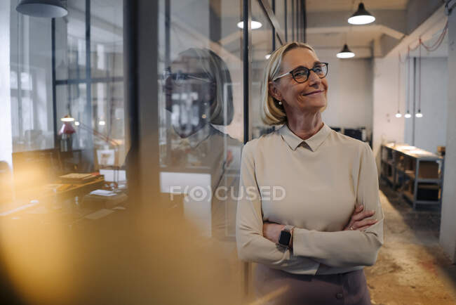 Porträt einer lächelnden, reifen Geschäftsfrau, die sich im Büro an eine Glasscheibe lehnt — Stockfoto