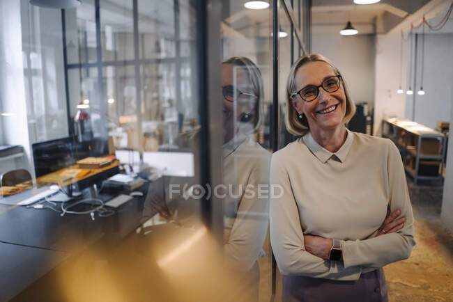 Porträt einer lächelnden, reifen Geschäftsfrau, die sich im Büro an eine Glasscheibe lehnt — Stockfoto