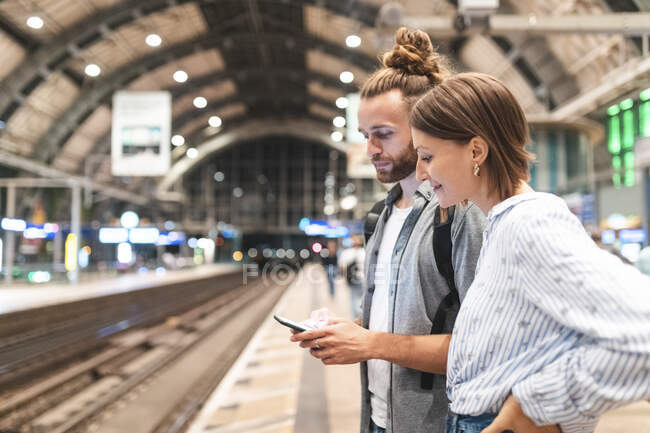 Jovem casal esperando o trem na estação e usando smartphone, Berlim, Alemanha — Fotografia de Stock
