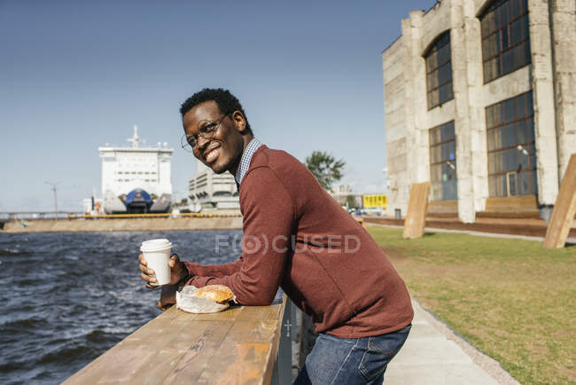 Joven mirando al mar, comiendo hamburguesa y café sobre barandilla - foto de stock