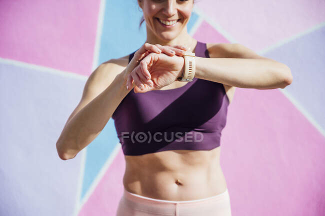 Усміхнена спортсменка дивиться на смарт-годинник, барвиста стіна на задньому плані — стокове фото