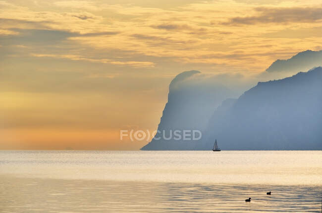 Italia, Trentino, Nago-Torbole, Silueta de velero navegando cerca de acantilados costeros del lago de Garda al amanecer - foto de stock