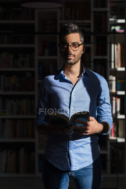 Porträt eines jungen Mannes mit Buch, der zu Hause vor Bücherregalen steht — Stockfoto