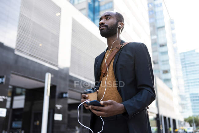 Giovane uomo d'affari con caffè per andare ad ascoltare musica con auricolari e smartphone in città — Foto stock