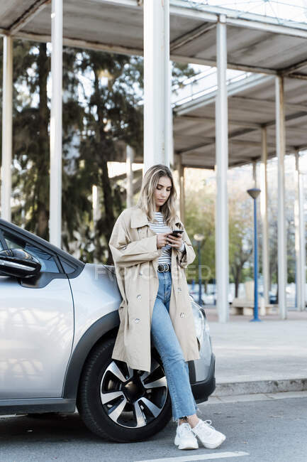 Mujer rubia joven usando un teléfono inteligente, apoyado en un coche - foto de stock