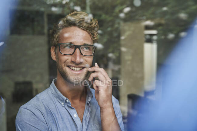 Lächelnder junger Geschäftsmann am Telefon hinter Fensterscheibe im Büro — Stockfoto