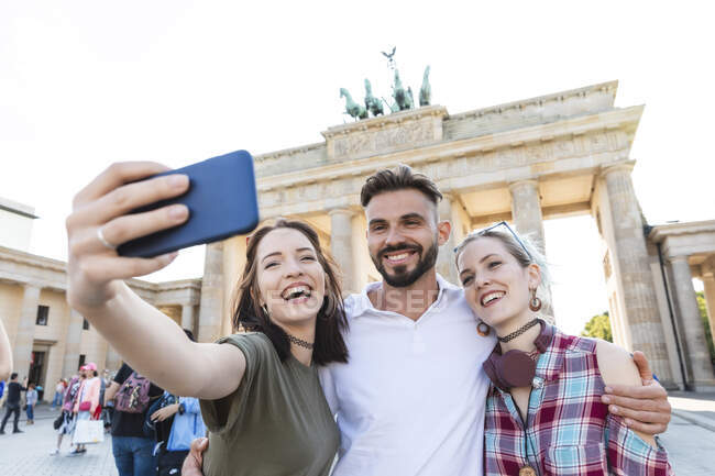Retrato de tres amigos felices tomando selfie con teléfono celular frente a Brandenburger Tor, Berlín, Alemania - foto de stock