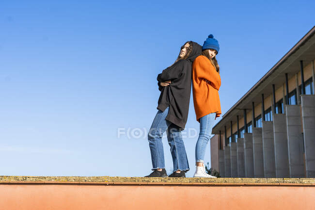 Two teenage girls standing back to back on a wall against blue sky — Stock Photo