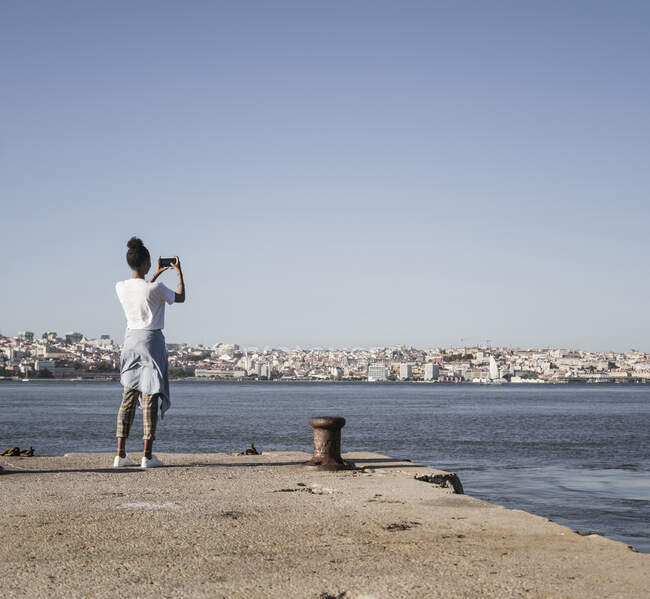 Jovem de pé no cais à beira-mar a tirar uma fotografia de telemóvel, Lisboa, Portugal — Fotografia de Stock