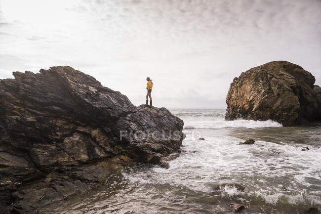 Mulher vestindo jaqueta de chuva amarela em pé na rocha na praia — Fotografia de Stock