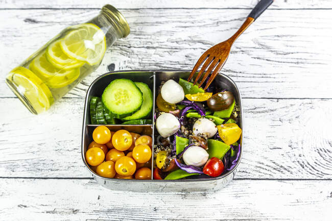Bottle of lemonade and lunchbox with cucumber slices, winter cherries and quinoa salad (quinoa, cherry tomato, red cabbage, sugar snap peas and mozzarella balls) — Stock Photo