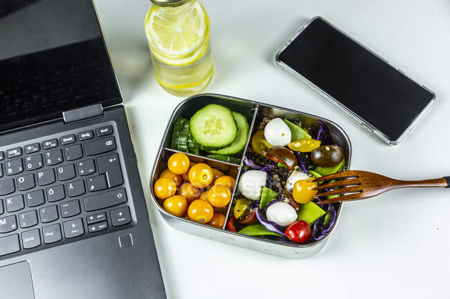 Laptop, smart phone, bottle of lemonade and lunchbox with cucumber slices, winter cherries and quinoa salad (quinoa, cherry tomato, red cabbage, sugar snap peas and mozzarella balls) — Stock Photo
