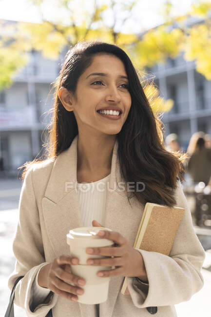Улыбающаяся молодая женщина с книгой и кофе на вынос в городе смотрит вокруг — стоковое фото