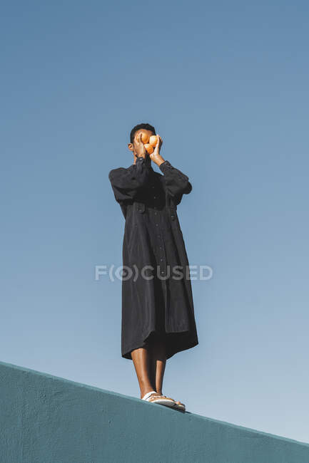 Joven vestido de kaftan negro parado en la pared azul sosteniendo naranjas frente a su cara - foto de stock