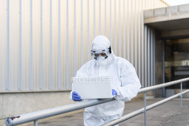 Female scientist wearing protective suit and mask and using laptop in front of the laboratory — Stock Photo