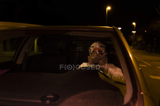 Senior woman wearing protective suit and mask driving car at night — Stock Photo