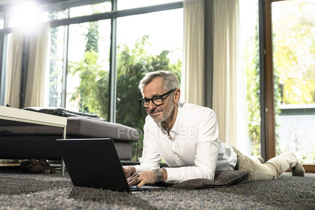 Sorrindo homem sênior com cabelos grisalhos na sala de estar de design moderno trabalhando no laptop — Fotografia de Stock