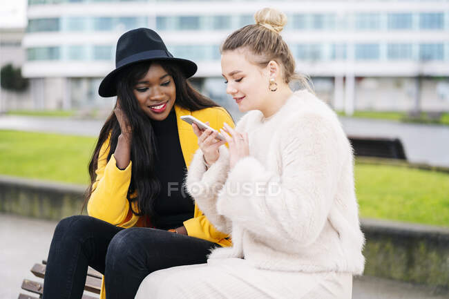 Девушки, сидящие на скамейке в городе с помощью мобильного телефона — стоковое фото