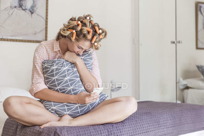Happy of young woman with curlers in hair sitting on bed looking at cell phone — Stock Photo