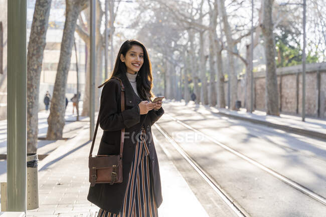 Portrait of smiling young woman with smartphone at the tram stop — Stock Photo
