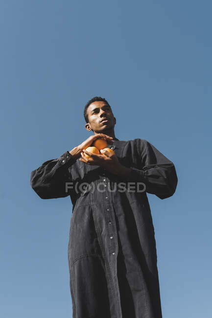 Молодой человек в черном кафтане под голубым небом держит апельсины — стоковое фото