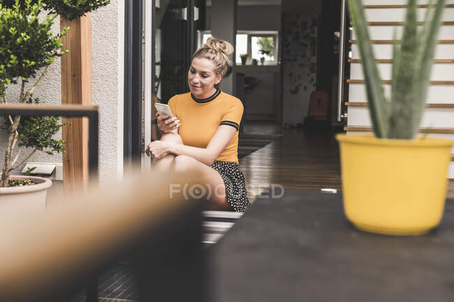 Donna rilassata seduta alla porta della terrazza a casa utilizzando il telefono cellulare — Foto stock