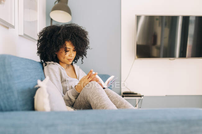 Mujer joven con el pelo rizado leyendo un libro en el sofá en casa - foto de stock
