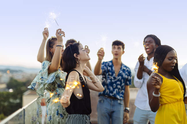 Group of happy multi-ethnic friends celebrating a party in the evening, holding sparklers — Stock Photo