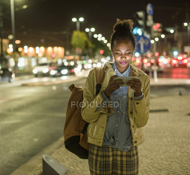 Retrato de mujer joven con auriculares y smartphone en la ciudad por la noche, Lisboa, Portugal - foto de stock