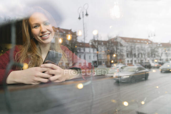 Portrait of happy young woman with mobile phone in a coffee shop looking out of window — Stock Photo