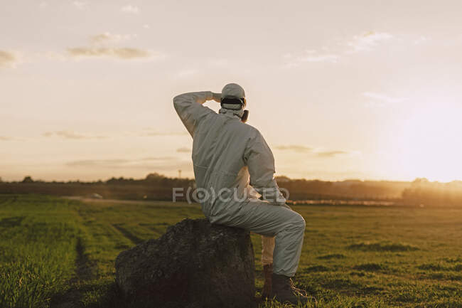 Hombre con traje protector sentado en una roca en el campo al atardecer - foto de stock